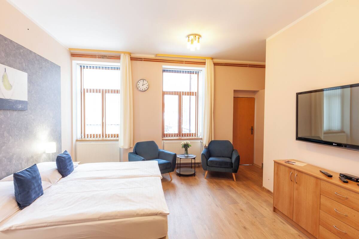 A hotel room with a bed, two armchairs, a small table, a TV, and a clock on the wall.