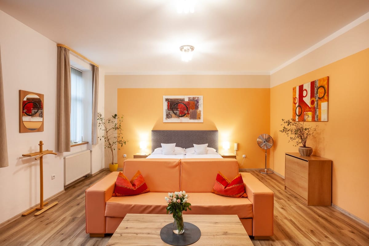 A hotel room with a bed, a sofa, a bedside table, a lamp, and a potted plant.