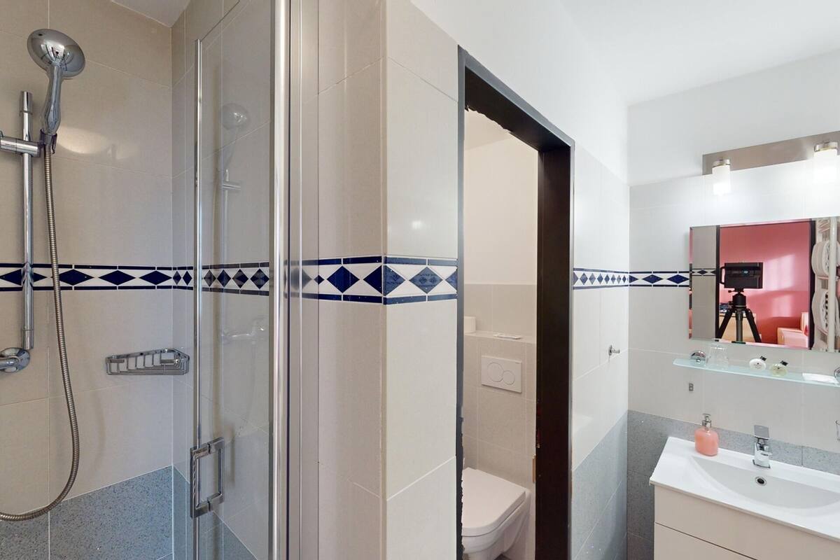 A modern bathroom with a glass shower enclosure, a toilet, a sink with a mirror, and a vanity with a pink soap dispenser.