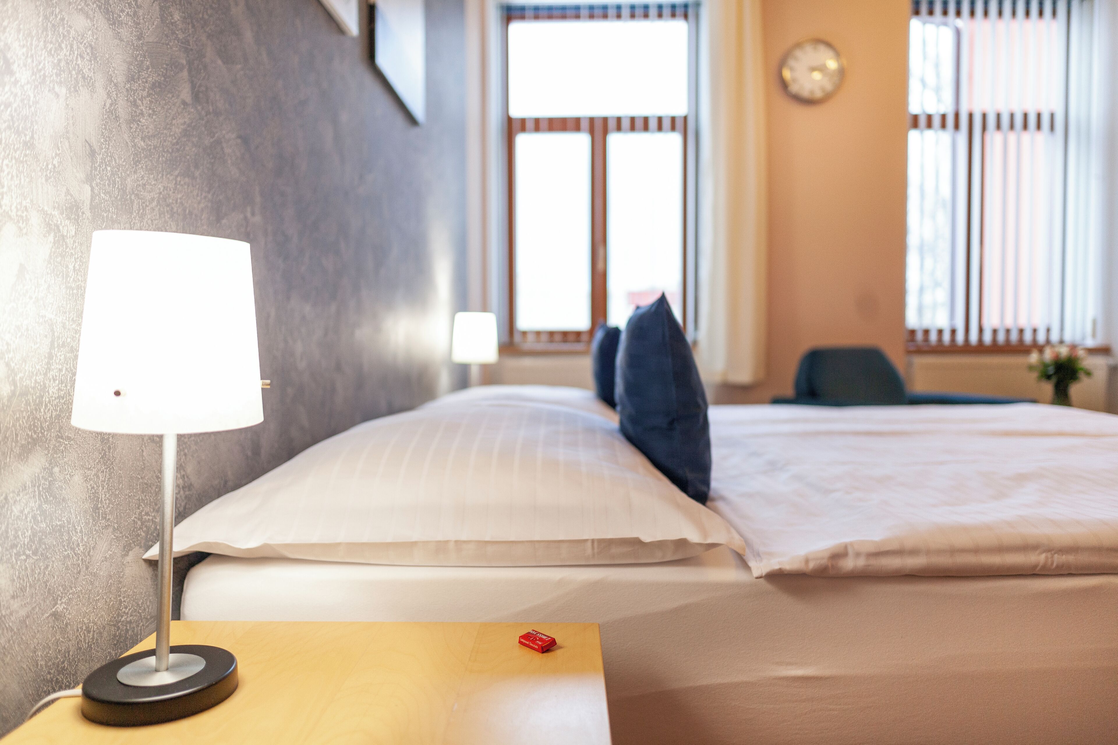 Une chambre d’hôtel avec un lit, une table de chevet avec une lampe, une fenêtre avec des stores et une horloge au mur.