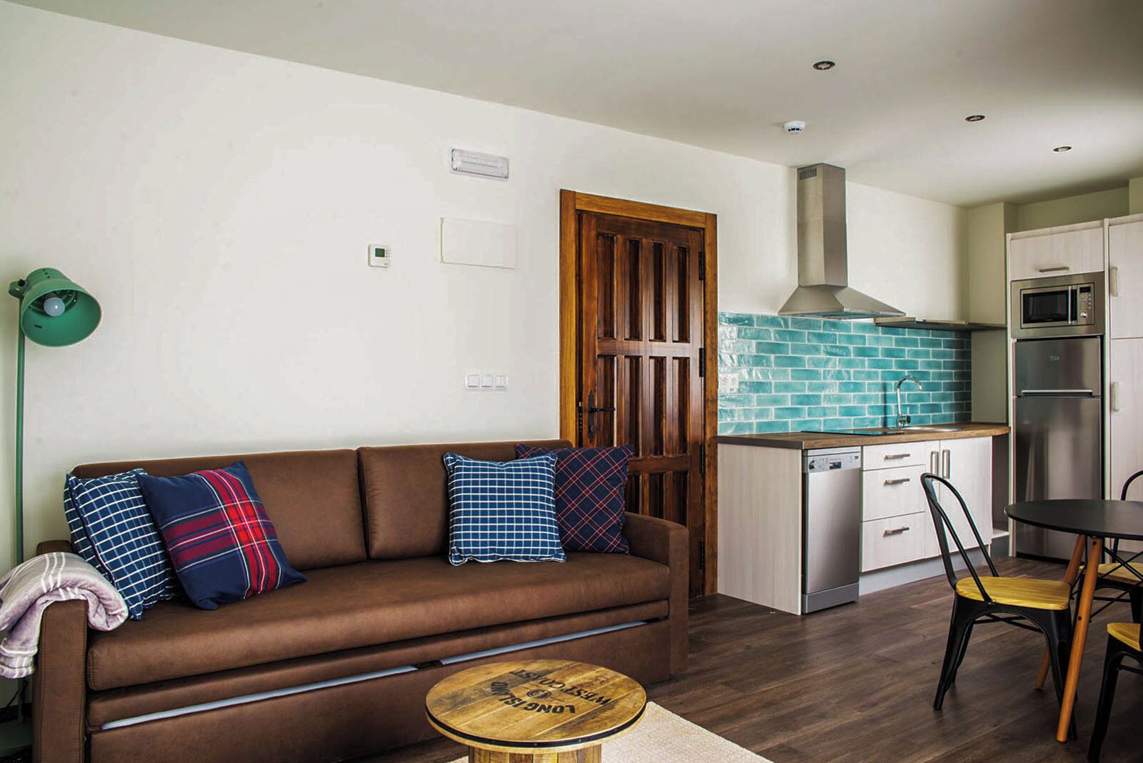 Ein modernes Wohnzimmer mit einer braunen Couch, karierten Kissen, einem kleinen Holztisch und einer Küchenzeile mit Mikrowelle, Backofen und Geschirrspüler.