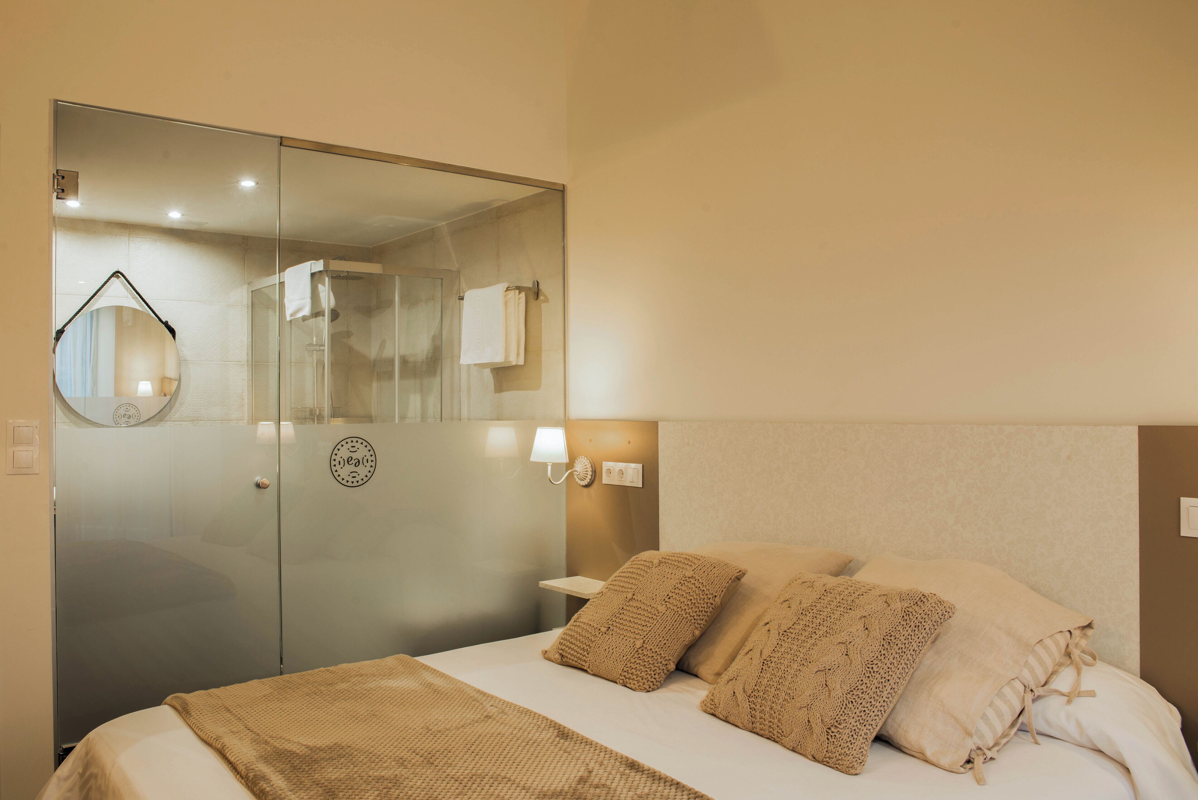 A hotel room with a bed, a glass shower enclosure, a round mirror, and a wall-mounted light fixture.