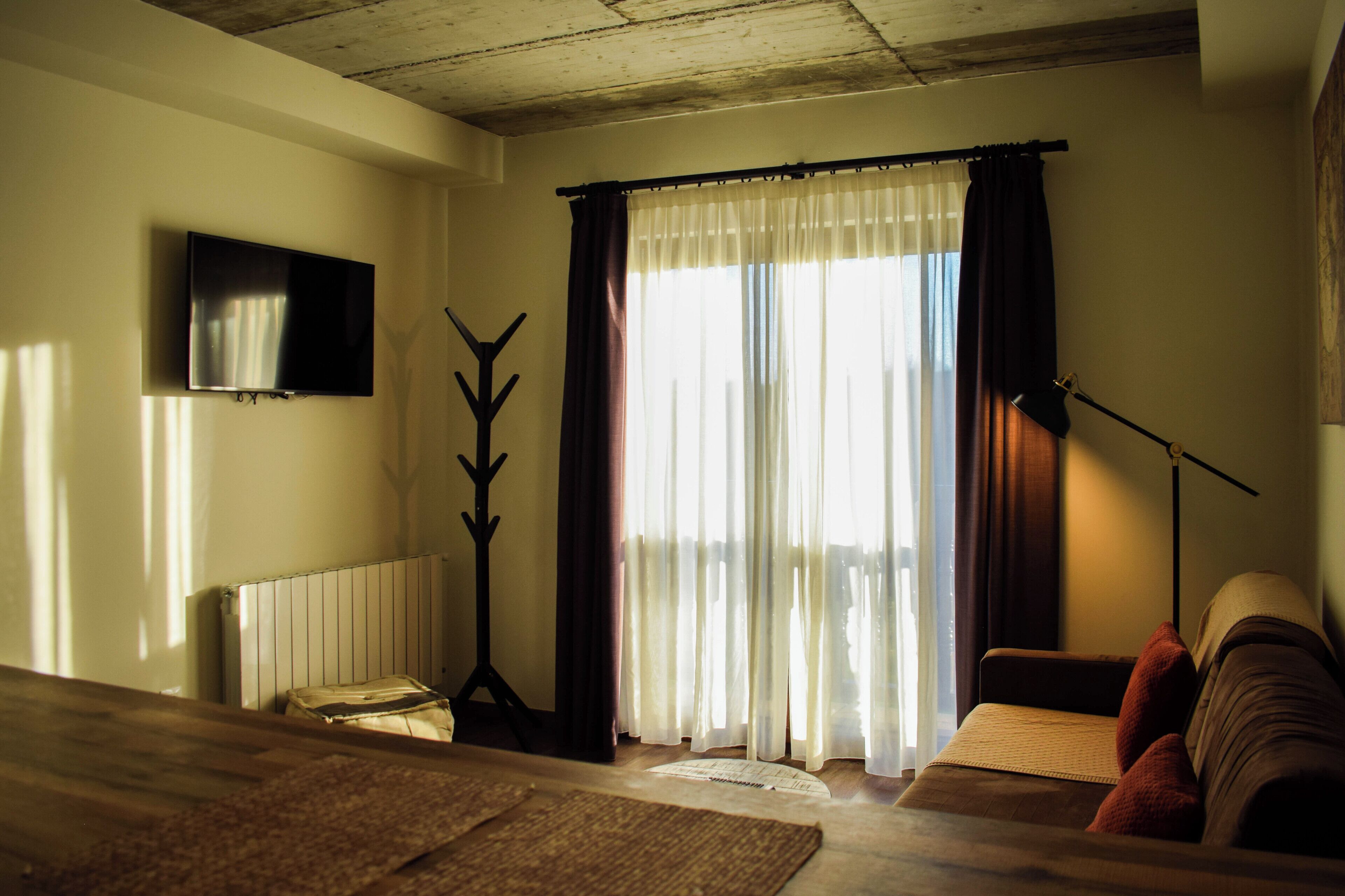 Un salon confortable avec un canapé, un téléviseur à écran plat fixé au mur, un radiateur et une grande fenêtre avec des rideaux.