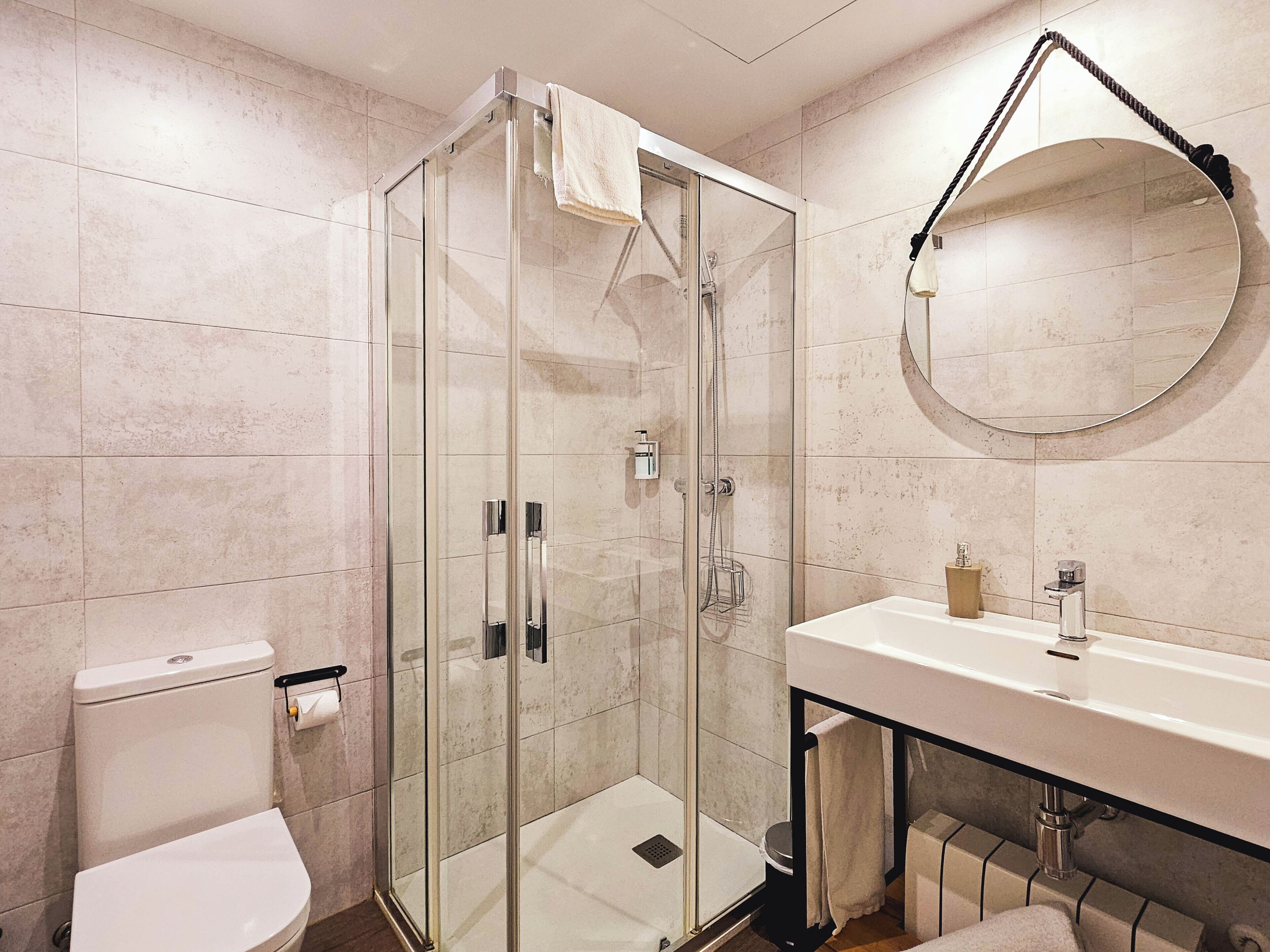 Un baño moderno con ducha envuelta en vidrio, espejo redondo, lavabo blanco y retrete.