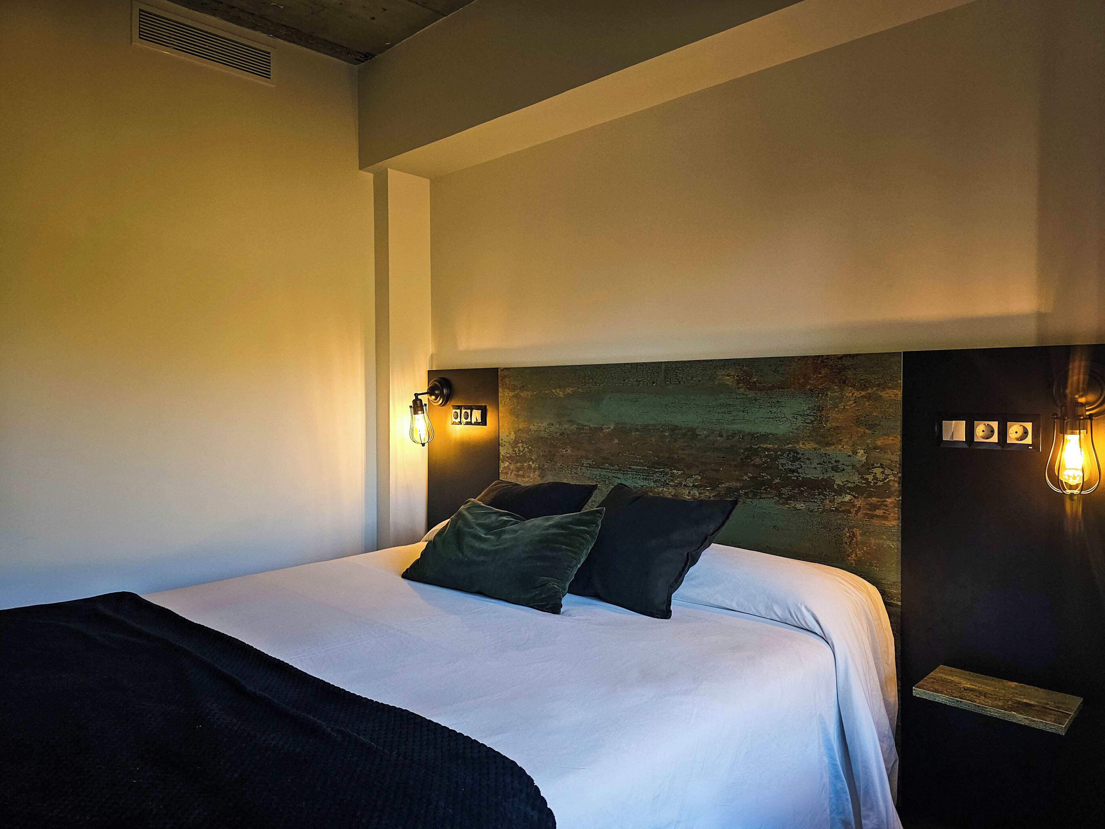 Un dormitorio moderno con una cama grande, dos lámparas de lectura fijadas a la pared y una pared rústica con textura detrás de la cama.