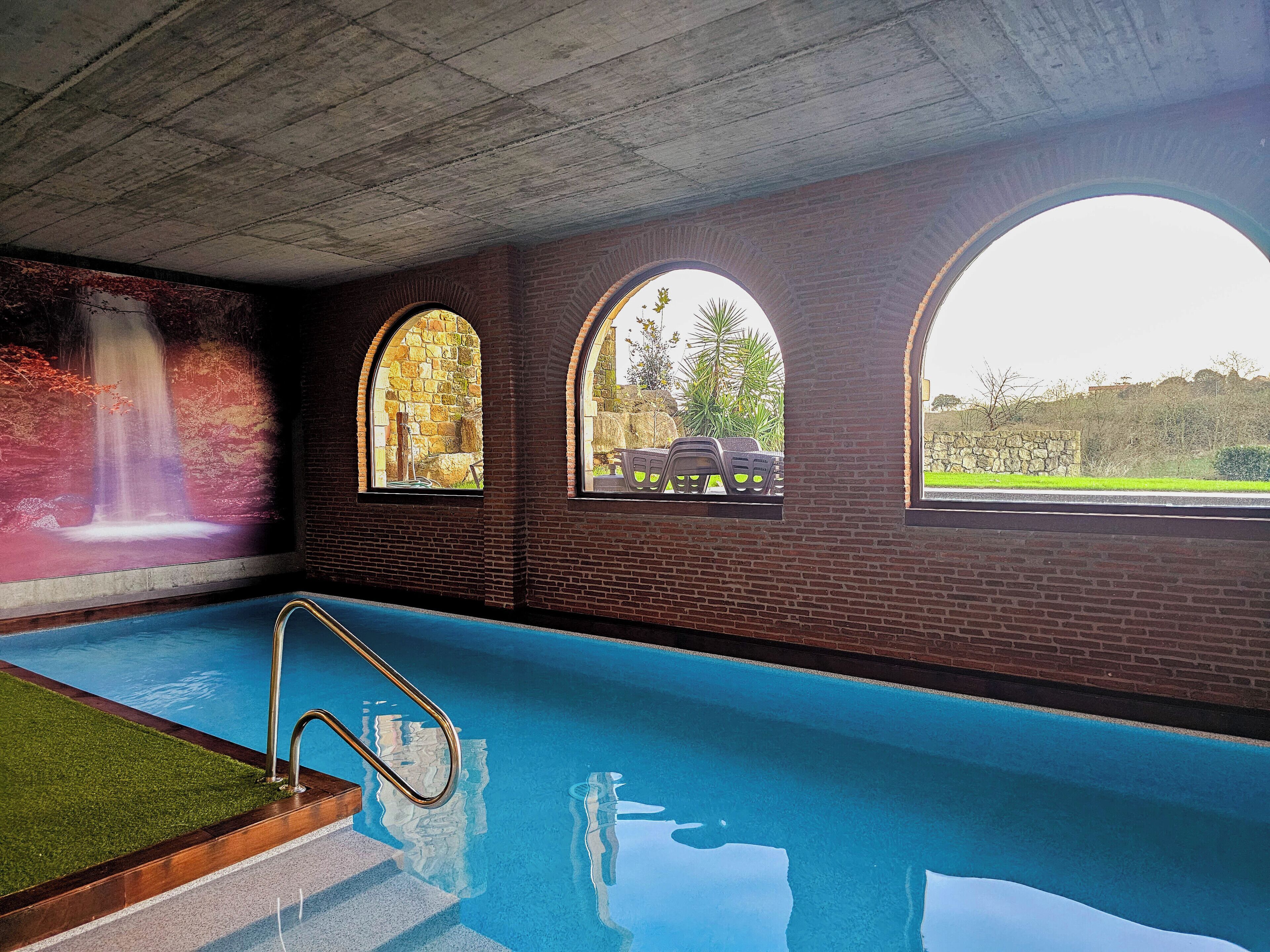 En indendørs swimmingpool med store glasvinduer, en metalstige og et muralmaleri af et vandfald.