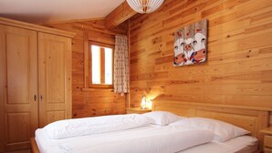 Una habitación de madera con cama, mesita de noche, ventana con cortinas y un cuadro en la pared.