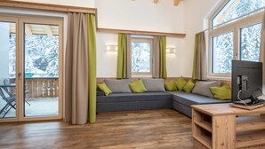 Een moderne woonkamer met een hoekbank, houten vloeren en grote ramen met uitzicht op besneeuwde bomen.