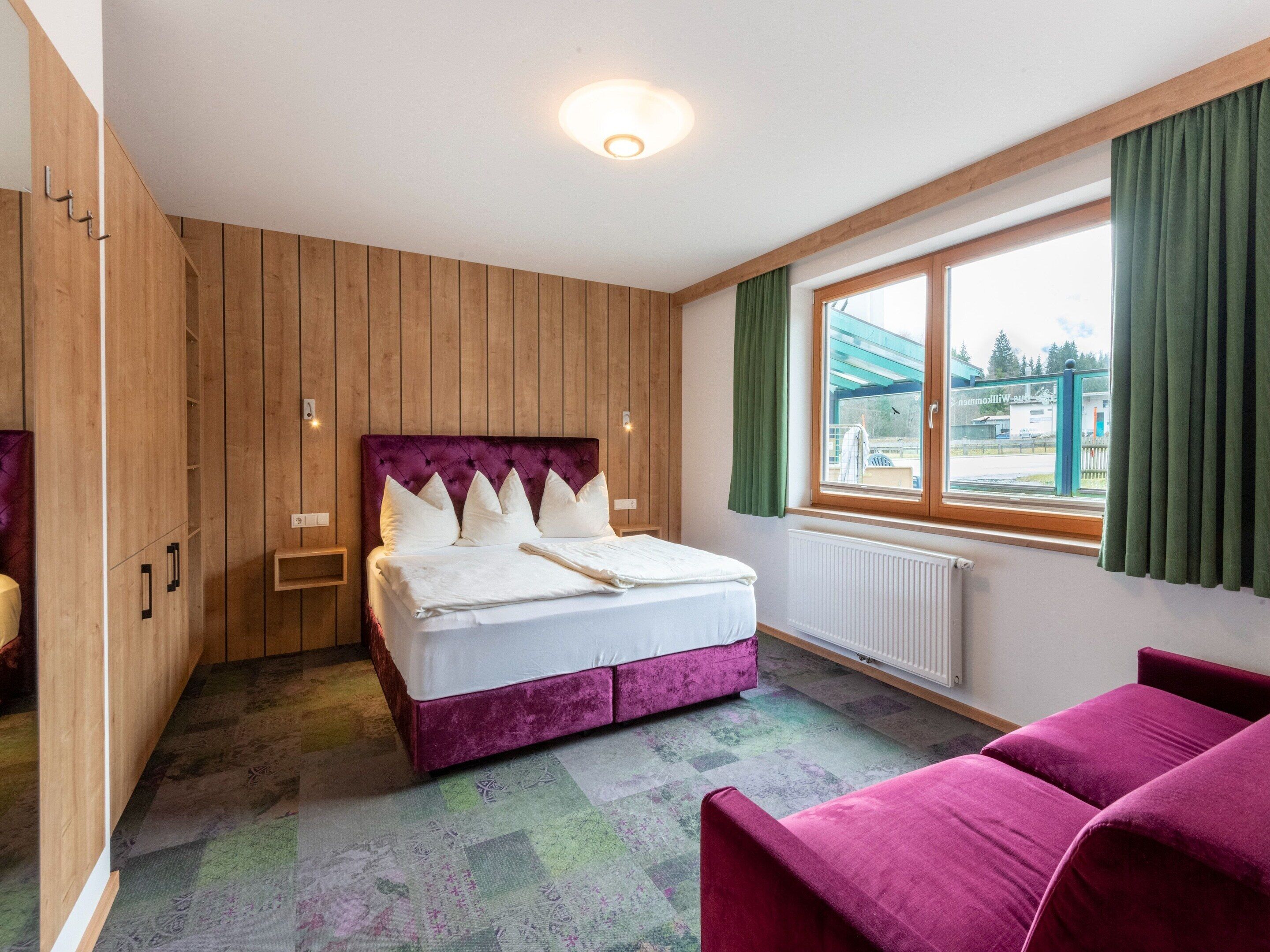 A bedroom with a bed, a window with curtains, a radiator, and a wooden wardrobe.