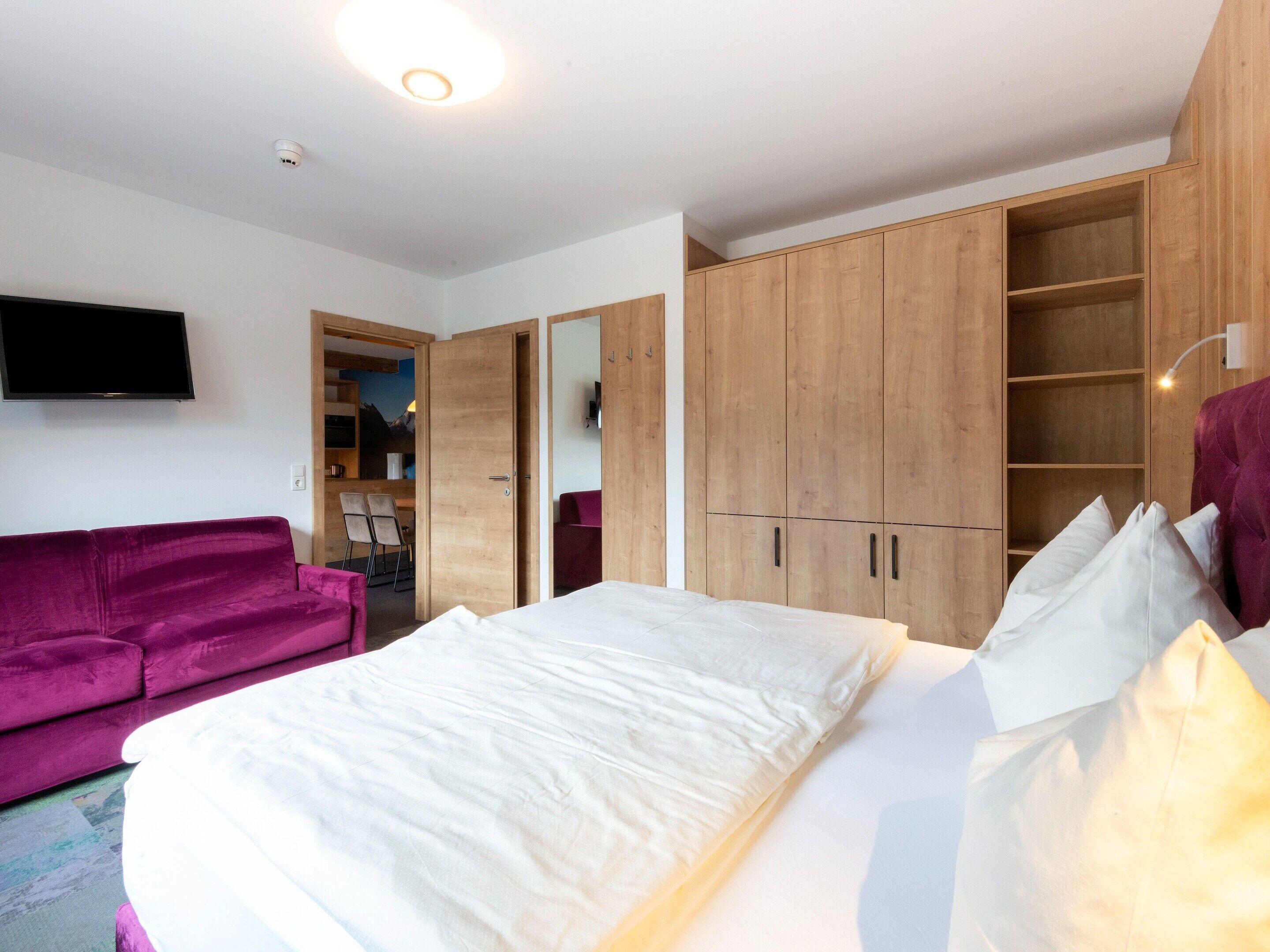 A bedroom with a bed, a purple sofa, wooden wardrobes, and a TV on the wall.