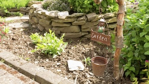 A rustic garden with a stone wall, a wooden sign reading "Garten", and a metal watering can hanging from a wooden post. - Ferienwohnung im Sauerland Hillside (Hallenberg)