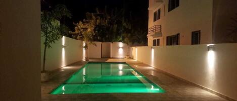 A rectangular swimming pool with a green-lit interior, surrounded by walls and potted plants, in a nighttime setting.