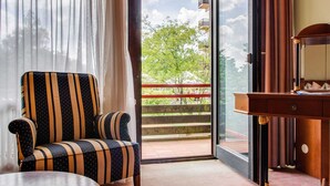 A room with a striped armchair, a wooden desk, and a view of a balcony with a railing and greenery.