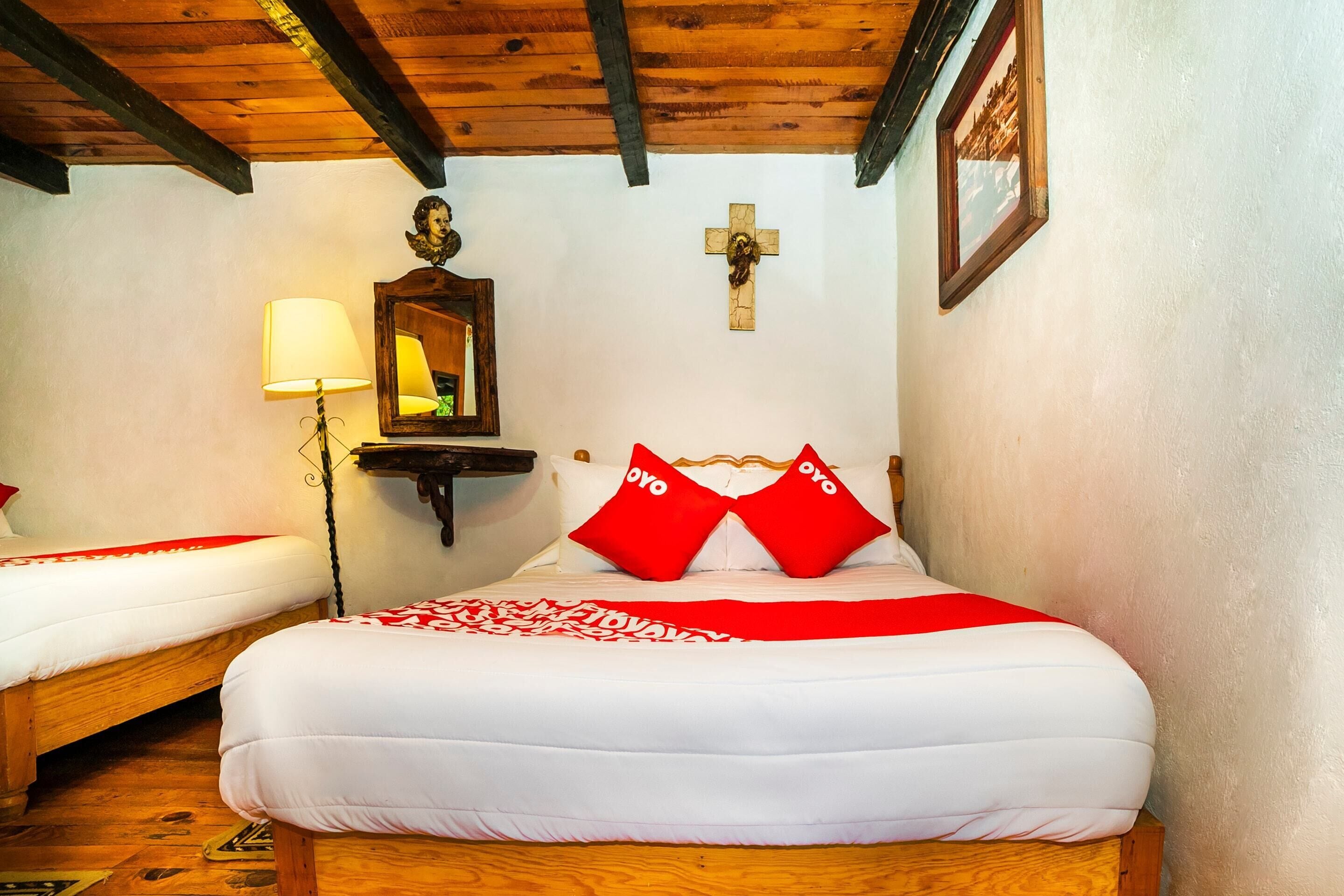 Un dormitorio con dos camas individuales, techo de madera, una cruz en la pared y un espejo sobre la cama izquierda.