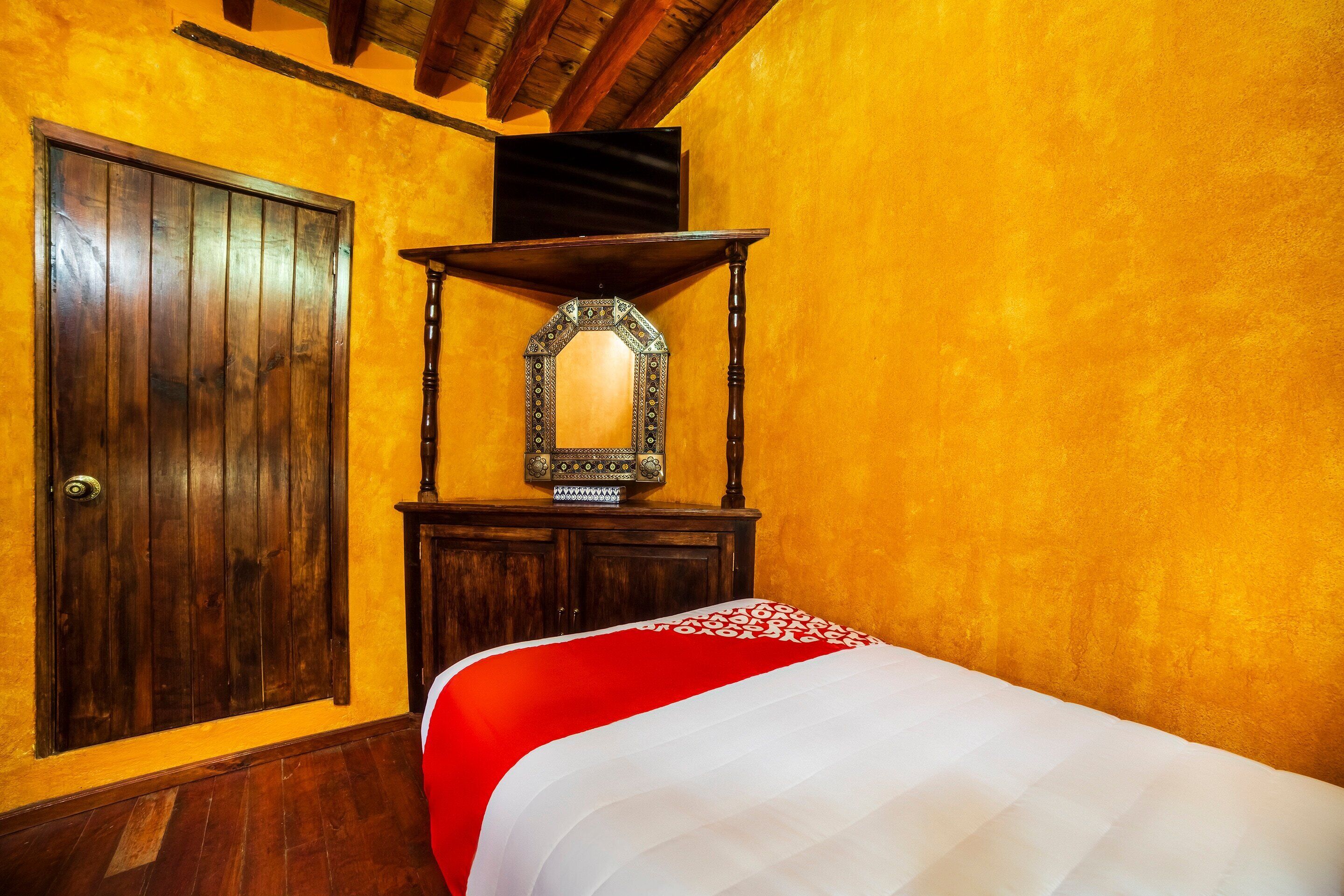 A room with a wooden door, a bed with a red and white bedspread, a wooden dresser, and a mirror on top of the dresser.