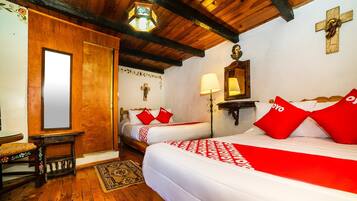 Quarto com vigas de madeira, duas camas com roupa de cama vermelha e branca, uma cadeira de madeira, uma mesa pequena e uma cruz na parede.