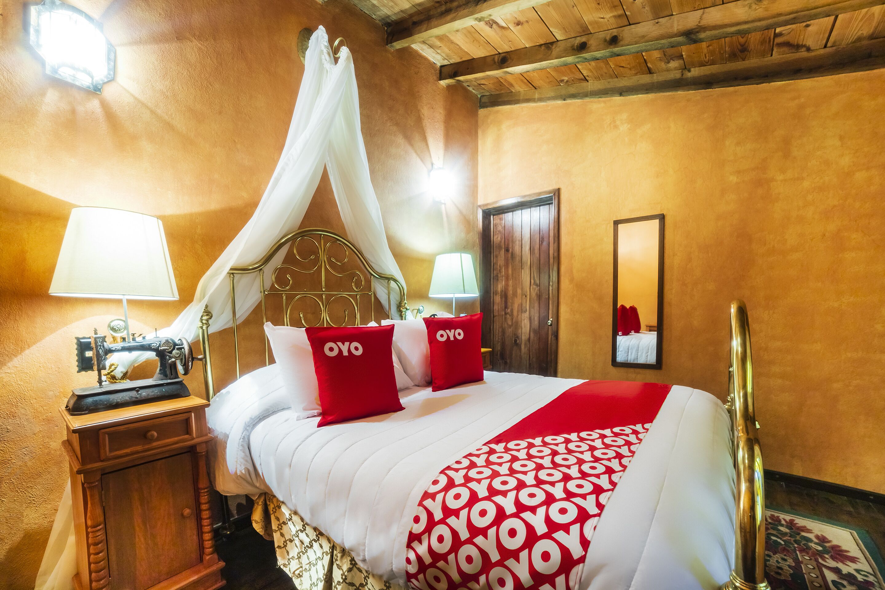 Quarto de hotel com cama de dossel, uma máquina de costura em uma mesinha lateral e um espelho na parede.