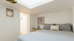 Un dormitorio con tragaluz, una cama con ropa de cama gris, una mesita de noche con una lámpara y un cuadro enmarcado en la pared.