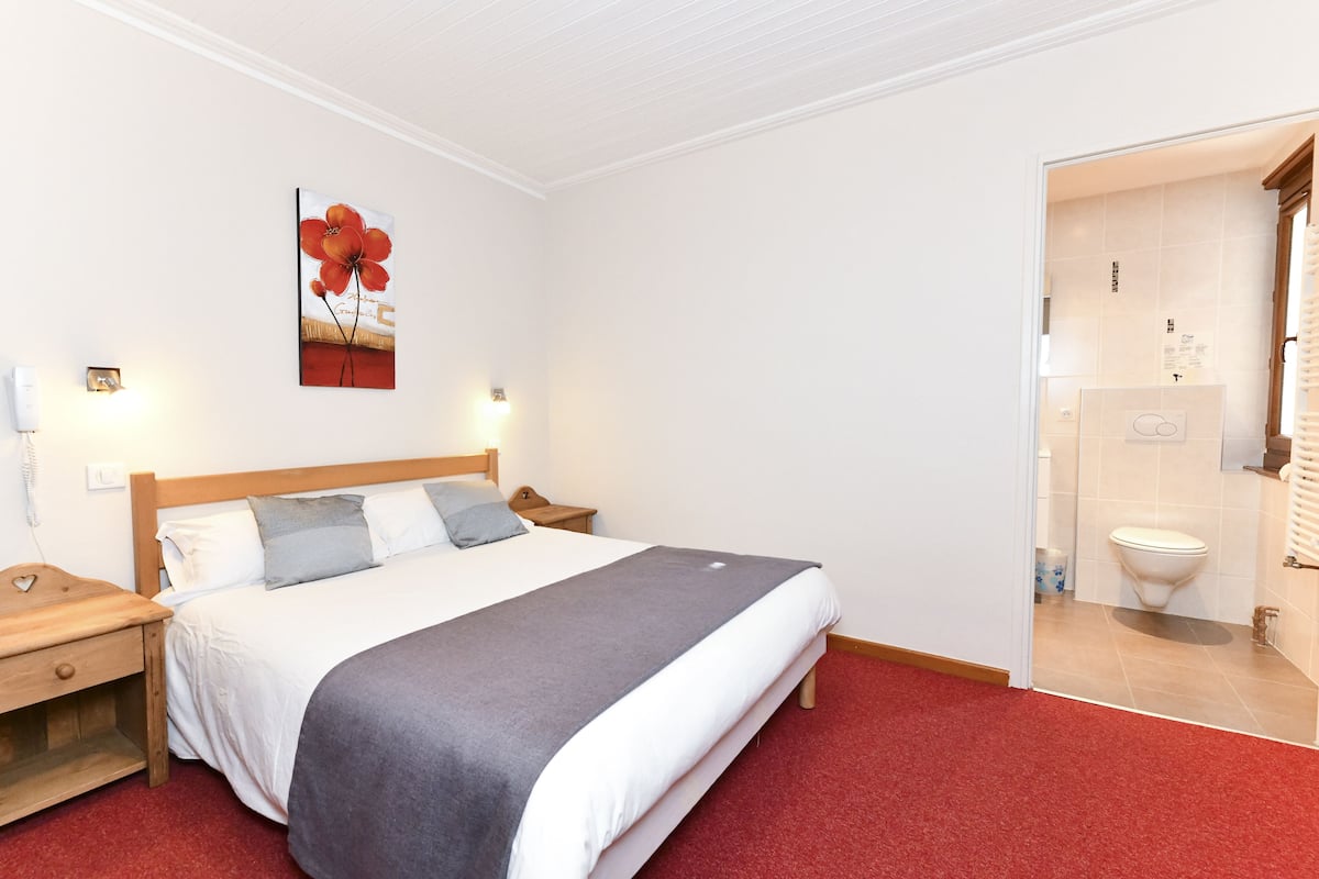 Una habitación de hotel con una cama grande, una mesita de noche, un cuadro en la pared y un baño visible a través de una puerta abierta.