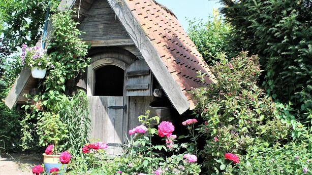 Eine rustikale Holzhütte mit geneigtem Dach, umgeben von üppigem Grün und blühenden rosa Blüten.