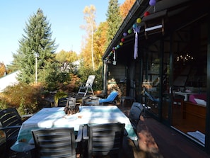Een buitenterras met een tafel en stoelen, omgeven door bomen en een gebouw met grote ramen.
