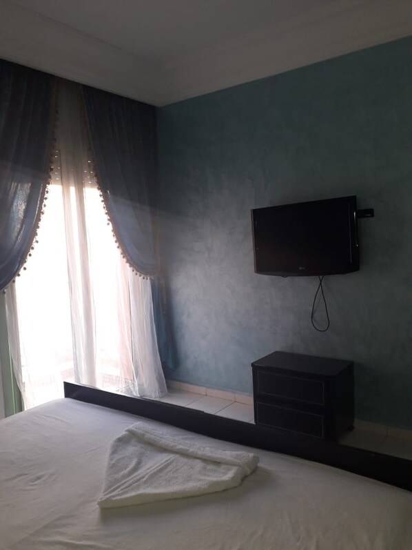 A bedroom with a bed, a flat-screen TV mounted on the wall, and a window with curtains.