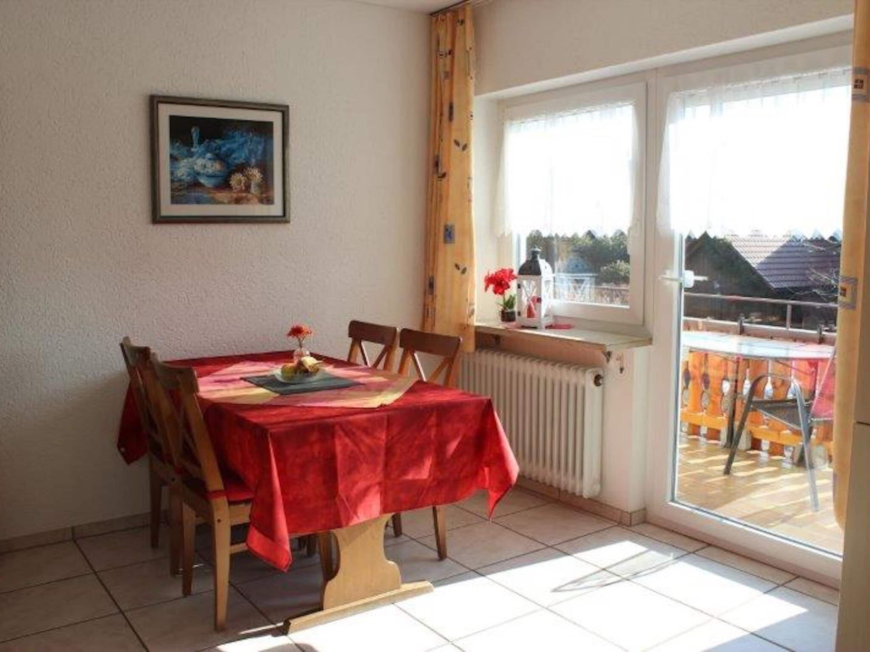 A dining area with a red tablecloth, wooden chairs, a framed painting, and a view through a window.