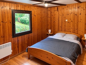 Une pièce aux murs lambrissés, un lit avec un matelas bleu, une fenêtre donnant sur la verdure et une table de chevet avec une lampe.