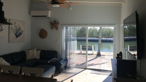 En stue med ventilator, sofa, bord og udsigt til et vandareal gennem store glasdøre.