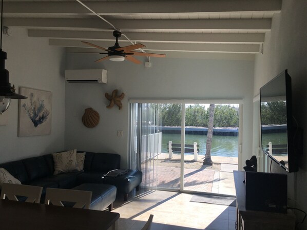 Una sala de estar con ventilador de techo, sofá, mesa y vista a un cuerpo de agua a través de puertas de vidrio grandes.