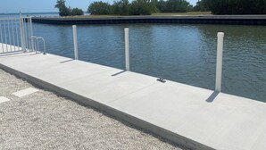En betonbro med gelændere og pæle, der stikker ud i stille vand, omgivet af træer og en klar himmel.