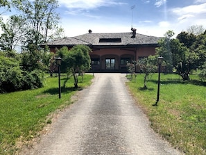 A building with a paved driveway leading to a gated entrance, surrounded by greenery and trees. - Villa Bonelli Bed & Fest (Savigliano)