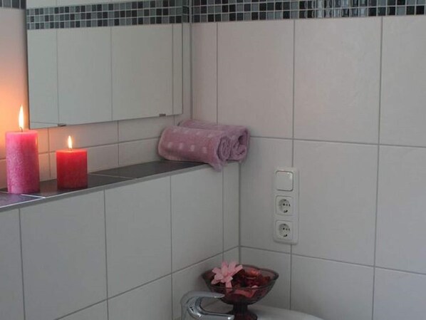A bathroom with white tiles, a mirror, two lit candles, a pink towel, and a glass bowl with a pink flower.