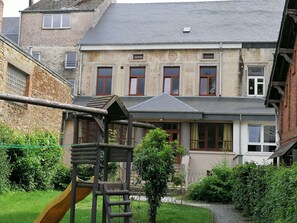 Un immeuble résidentiel à la façade en briques, doté de nombreuses fenêtres et d’un petit espace jardin agrémenté d’une structure de jeux en bois.