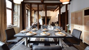Un coin repas avec une longue table dressée pour six personnes, des poutres en bois au plafond, des murs en pierre et, au fond, une vue sur la cuisine.