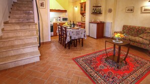 Un escalier mène à un coin repas où une table est dressée pour deux personnes. On aperçoit une petite cuisine au fond, et un canapé avec un panier de fruits.