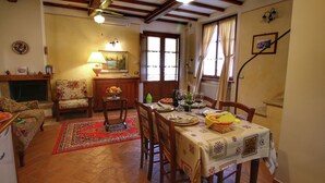 Un coin repas avec une table en bois dressée pour quatre personnes, entourée de chaises. On y trouve une cheminée avec un manteau de cheminée, une lampe et un tableau accroché au mur.