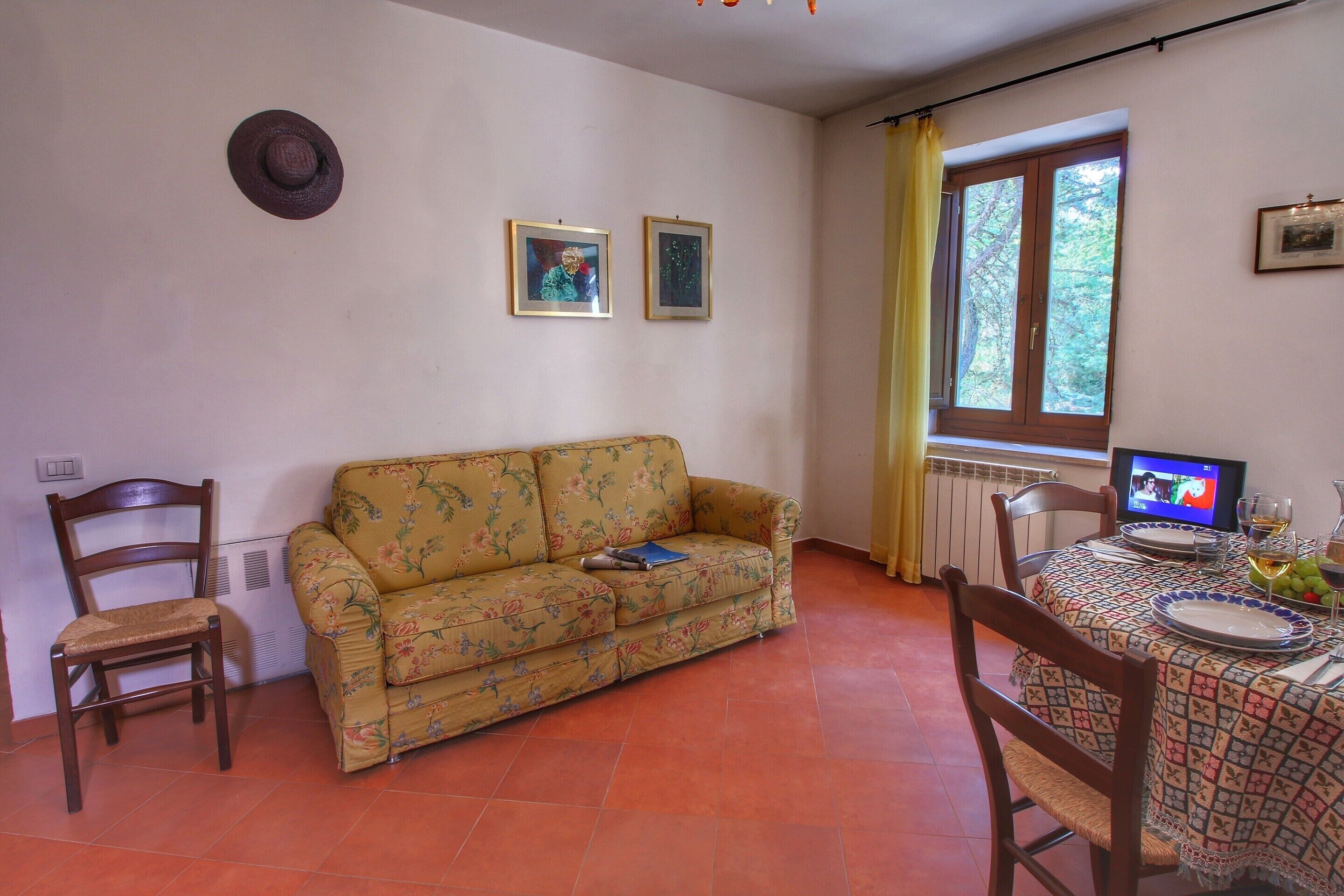 Ein Wohnzimmer mit einer Couch, Sesseln, einem Esstisch mit Tischdecke, einem Fernseher und einem Fenster mit Vorhängen.