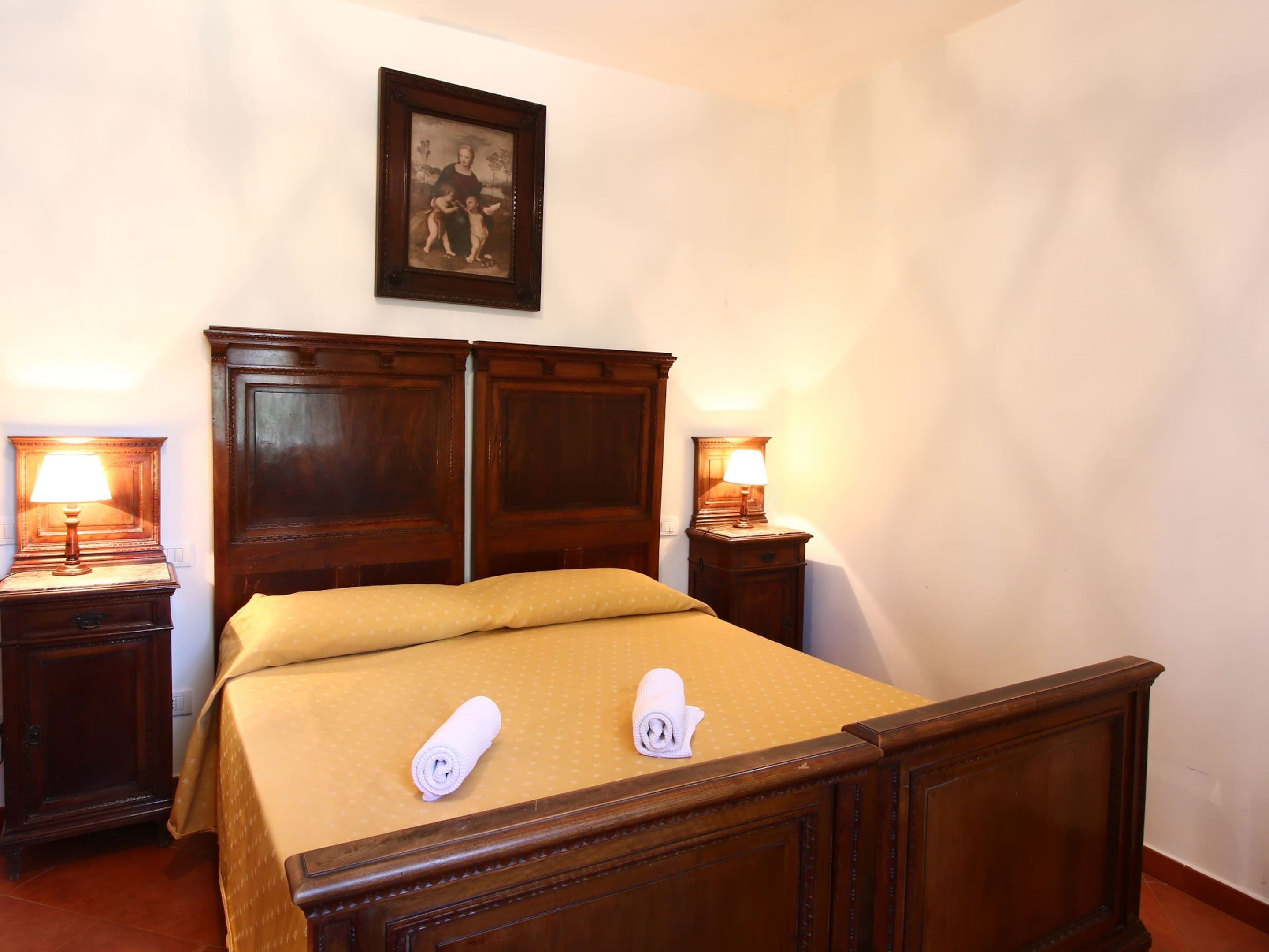 A hotel room with a wooden bed, two bedside tables with lamps, a framed picture on the wall, and two towels on the bed.