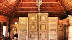 A room with a high wooden ceiling, a large ornate partition with intricate designs, a hanging light fixture, and a wooden chair. -  Mimosa chalet Indonésie. (Essaouira)