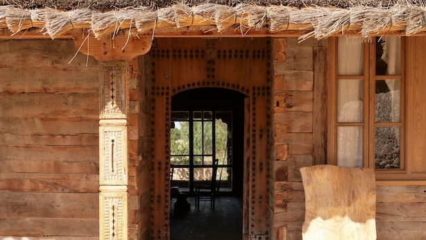 A wooden building with a thatched roof entrance, a large wooden door, and a window with a stone wall behind it. -  Mimosa chalet Indonésie. (Essaouira)