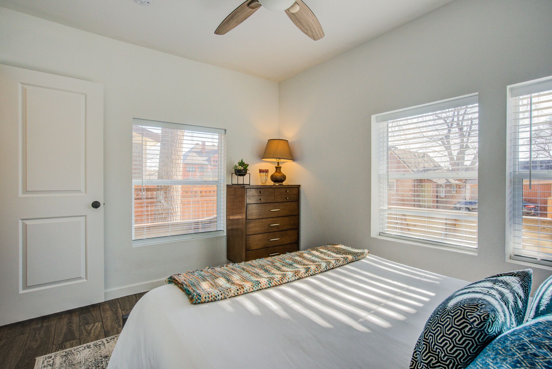 A bedroom with a bed, a dresser, a lamp, and two windows with blinds.