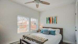 Een slaapkamer met een bed, een plafondventilator, een raam met jaloezieën en een kunstwerk aan de muur.