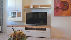 A modern living room with a flat-screen TV mounted on a wooden cabinet, a small shelf above the TV, and a large floral painting on the wall.