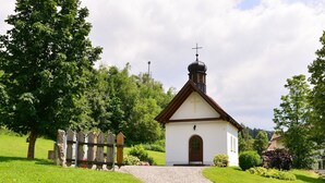 A small white church with a bell tower and a cross on top, surrounded by greenery and a stone fence. - Ferienhaus mit Sauna in Skigebietsnahe (Drachselsried)