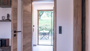 An open wooden door leading to a patio with a table and chairs, a bookshelf with books, and a small speaker on a shelf. - Ferienhaus mit Sauna in Skigebietsnahe (Drachselsried)