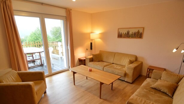 Een woonkamer met een bank, fauteuil, salontafel en een lamp. Er is een schuifdeur die leidt naar een buitenruimte.
