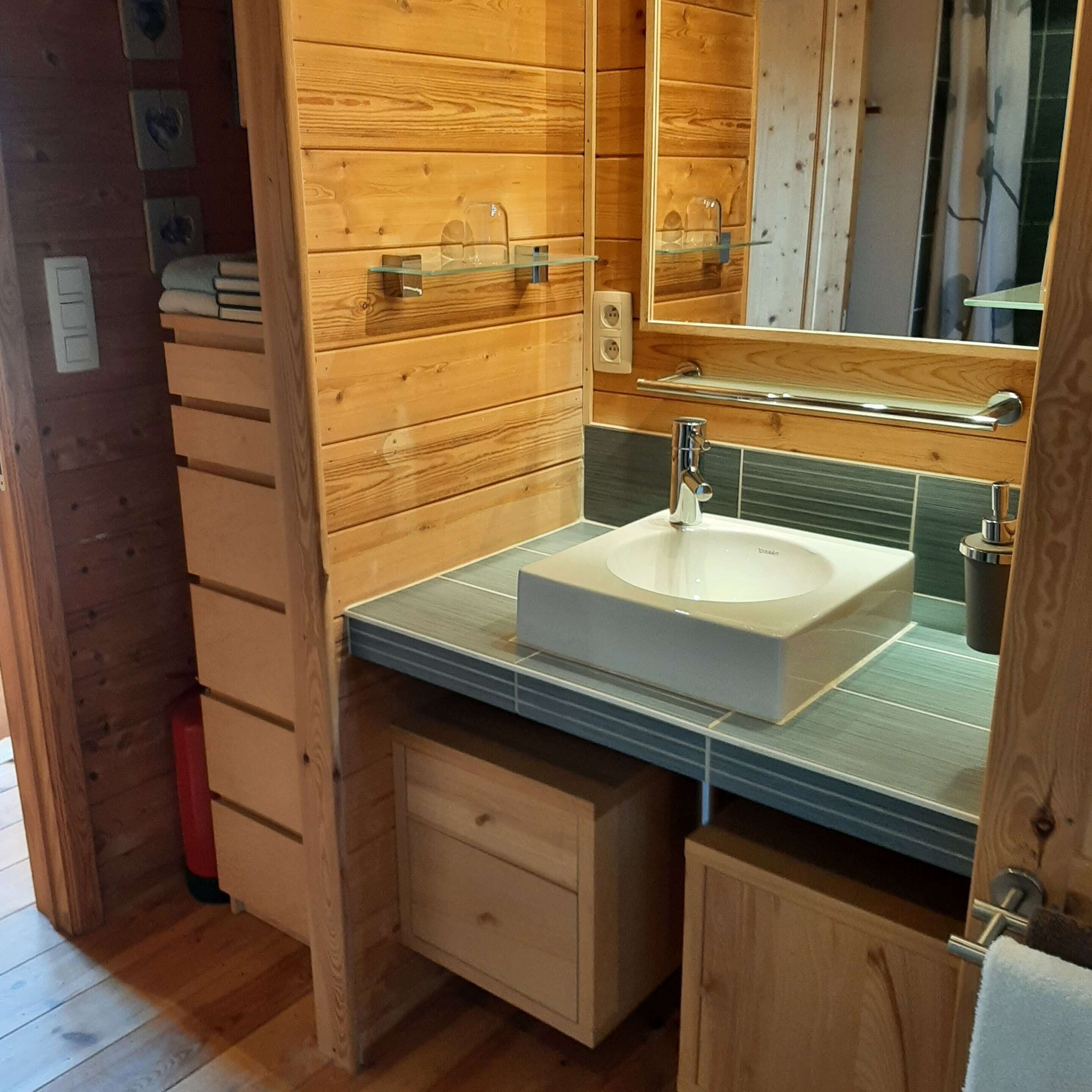 Une salle de bain aux murs lambrissés, avec un lavabo blanc, un miroir et un petit meuble.