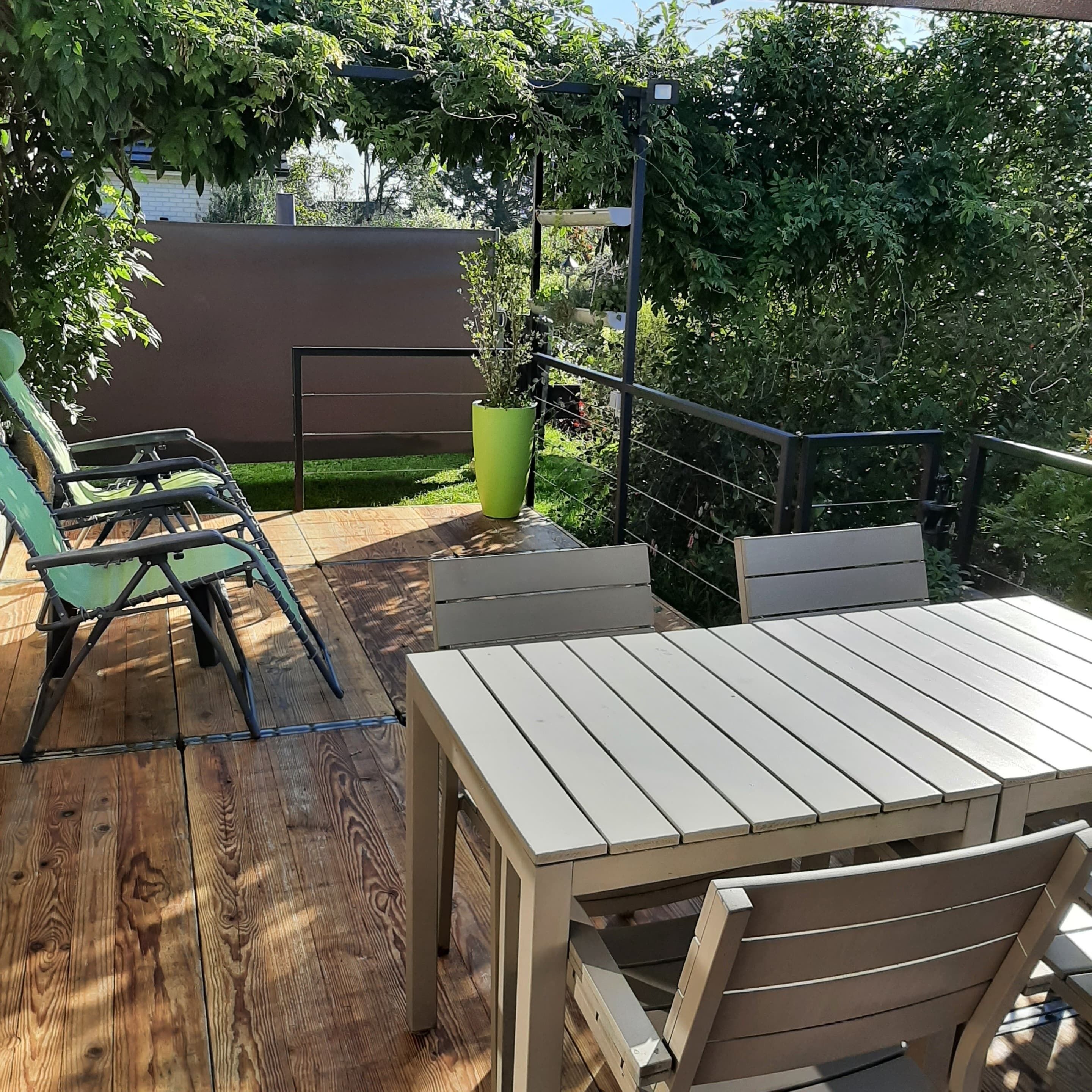A wooden deck with a table and chairs, surrounded by greenery and a metal railing.