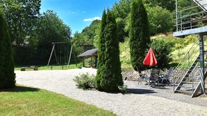 Un chemin de gravier mène à un kiosque, entouré de verdure et surmonté d’un parasol rouge.